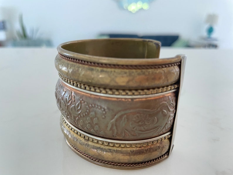 Puede incluir: Pulsera de pu&ntilde;o ancha con acabado de lat&oacute;n antiguo. La pulsera presenta tres bandas apiladas con intrincados patrones florales en relieve y trabajos decorativos en metal. El pu&ntilde;o mide aproximadamente 6,35 cm de ancho.