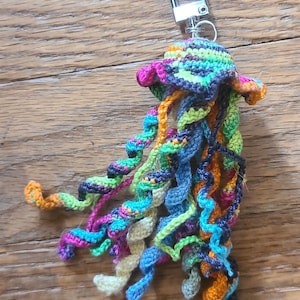 May include: A colorful, handmade crochet jellyfish keychain. The jellyfish features a rainbow of colors, including pink, green, blue, and orange. It has a silver-colored metal clasp for attaching to keys or bags.