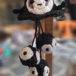 May include: A handmade crochet charm featuring a No-Face character from Spirited Away, with a white face, purple accents, and black body. Below, three smaller black and white charms dangle from black strings. The charm is suspended from a wooden surface.