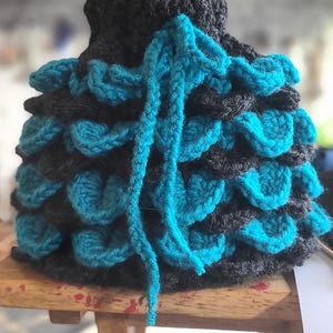 May include: A handmade crochet item, possibly a hat or cowl, in shades of teal and charcoal gray. The design features a layered, scale-like pattern with a long teal tie. The item is displayed on a small wooden surface.