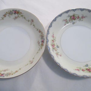 May include: Two white porcelain bowls with floral patterns. One bowl has a gold rim and a floral garland. The other bowl has a blue patterned rim and floral accents. Both bowls have a white centre.