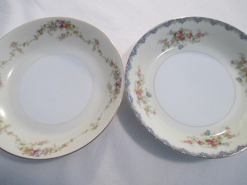 May include: Two white porcelain bowls with floral patterns. One bowl has a gold rim and a floral garland. The other bowl has a blue patterned rim and floral accents. Both bowls have a white centre.