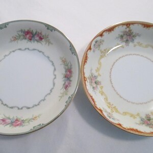 May include: Two decorative porcelain bowls with floral designs. One bowl has a cream background with pink and blue flowers and a green border. The other bowl has a cream background with pink flowers and a gold and orange border.
