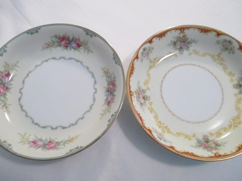 May include: Two decorative porcelain bowls with floral designs. One bowl has a cream background with pink and blue flowers and a green border. The other bowl has a cream background with pink flowers and a gold and orange border.