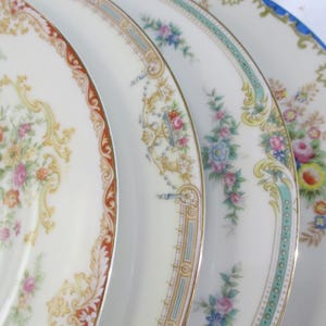 May include: A set of four stacked ceramic plates featuring cream-colored surfaces with distinct decorative borders. Each plate displays unique floral patterns in shades of pink, yellow, and blue, accented by intricate gold, red, and blue trim.