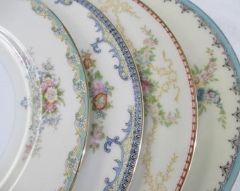 May include: Four ceramic plates arranged in a row, each featuring a unique decorative rim. The designs include floral patterns in pink, blue, and green, intricate scrollwork, and geometric borders in red, gold, and black on a cream-colored base.