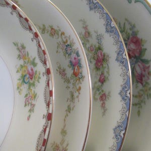 May include: Close-up of four decorative plates with floral patterns. The plates have a cream background and gold rims. Each plate features a different floral design in pink, blue, yellow, and green.