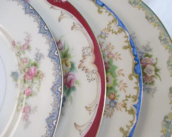 May include: Four ceramic dinner plates arranged in a row, each featuring a unique decorative border. The designs include floral patterns in pink, blue, and green, with accents of red, gold, and blue trim along the scalloped or smooth edges.
