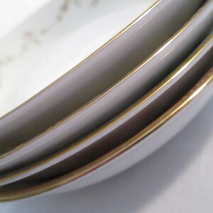 May include: Close-up of a stack of white porcelain dishes with a gold rim. The dishes have a slight oval shape and a delicate floral pattern in the upper left corner. The gold rim adds a touch of elegance to the tableware.