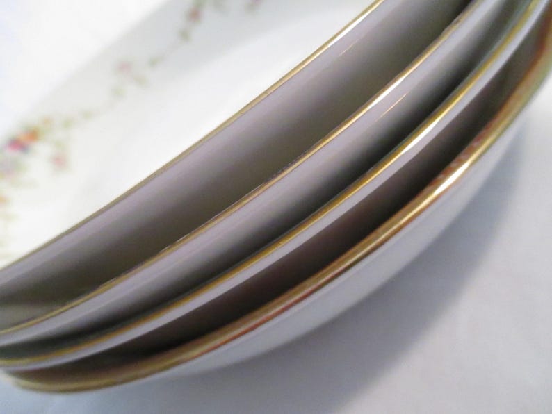 May include: Close-up of a stack of white porcelain dishes with a gold rim. The dishes have a slight oval shape and a delicate floral pattern in the upper left corner. The gold rim adds a touch of elegance to the tableware.