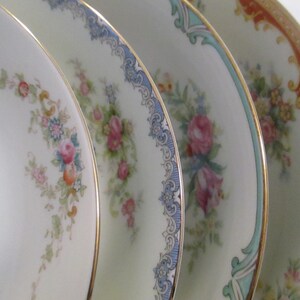May include: Close-up of four antique porcelain plates. Each plate has a different floral design with gold rims. The plates feature pink, blue, and orange colours, set against a white background. The plates are likely vintage.