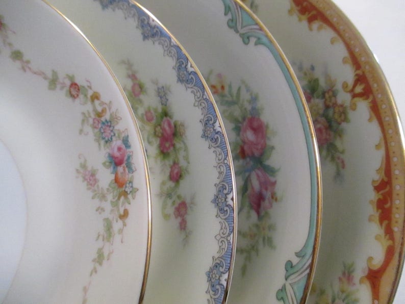 May include: Close-up of four antique porcelain plates. Each plate has a different floral design with gold rims. The plates feature pink, blue, and orange colours, set against a white background. The plates are likely vintage.