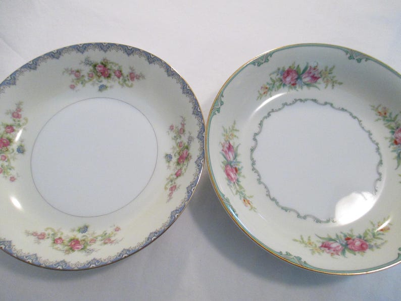 May include: Two white porcelain bowls with floral patterns. One has a blue and gold rim with pink and blue flowers. The other has a gold rim with pink flowers and a green border. Both bowls have a white centre.