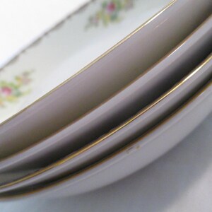 May include: A stack of white oval serving dishes with a gold trim. The dishes feature a floral pattern in pink, yellow, and green on the rim. The edges of the dishes are slightly curved, and the gold trim adds a touch of elegance.