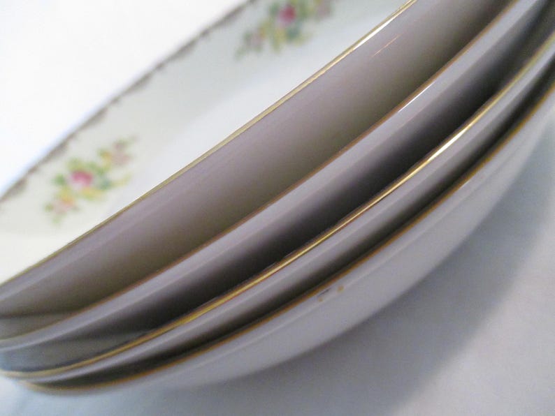 May include: A stack of white oval serving dishes with a gold trim. The dishes feature a floral pattern in pink, yellow, and green on the rim. The edges of the dishes are slightly curved, and the gold trim adds a touch of elegance.