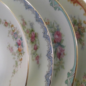 May include: Close-up of four antique porcelain plates with floral patterns. Each plate has a different design, including roses and other flowers, with gold rims. The plates are in shades of white and cream.