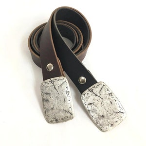 Puede incluir: Dos cinturones de cuero, uno marrón y otro negro, con hebillas rectangulares plateadas. Las hebillas tienen un aspecto texturizado y envejecido. Cada cinturón está asegurado con un remache plateado. Los cinturones se muestran sobre un fondo blanco.