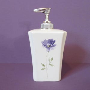 May include: A white ceramic soap dispenser with a tapered, rectangular body. The front features a painted purple flower with green leaves and a stem. It is topped with a silver-colored metallic pump mechanism with a clear plastic collar.