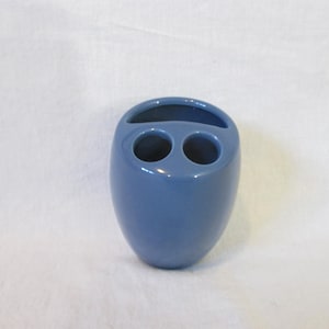 May include: A smooth, glossy, slate blue ceramic toothbrush holder with a rounded, egg-like shape. The top surface features three distinct openings: one elongated, curved slot and two circular holes designed for holding oral care items.