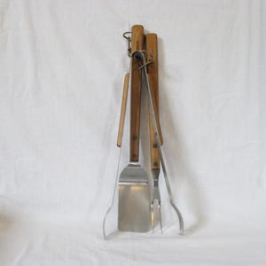 May include: A set of grilling tools, including a spatula and fork, with wooden handles and stainless steel heads. The tools are attached to a wooden holder with a metal loop for hanging. The set is designed for outdoor cooking.