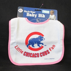 May include: White baby bib with a pink trim, featuring a blue bear logo inside a red circle, and the text "Little Chicago Cubs Fan". The bib has a tag that reads "McArthur Baby Bib".