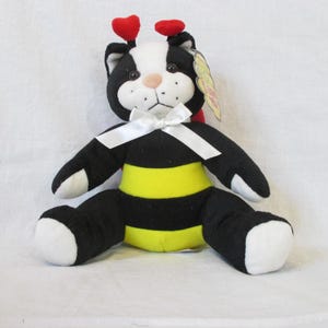 May include: A plush stuffed animal designed as a cat wearing a black and yellow striped bee costume. The cat has a white face, black ears with red heart-shaped antennae, white paws, and a white ribbon tied around its neck. A tag is attached to the ear.