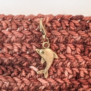 May include: A gold-toned dolphin charm attached to a crochet stitch marker. The marker is on a background of red crochet fabric.
