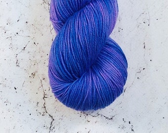 Può includere: Un gomitolo di filato tinto a mano nei toni del blu e del viola. Il filato è avvolto strettamente, mostrando una sfumatura di colori dall'indaco scuro al viola più chiaro. Il filato è esposto su uno sfondo bianco strutturato.