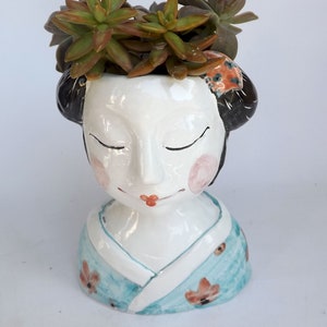 以下が含まれることがあります： 白い陶器製の植木鉢で、芸者の顔の形をしており、目を閉じて頬がピンク色です。芸者は、花のアクセントが付いた青い着物を着ています。植木鉢には、緑と茶色の多肉植物が植えられています。