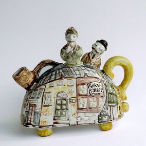Può includere: Teiera decorativa in ceramica con un design a scena di strada che include il testo "Baker Street" e il numero "221B". Due piccole figure di uomini sono in cima. La teiera ha un manico e piedini gialli.