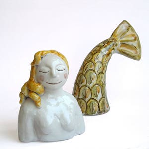 Pode incluir: Uma escultura de cerâmica de uma sereia com os olhos fechados e uma expressão serena. A sereia tem cabelo loiro e um corpo azul claro. Uma cauda curva separada em tons de verde e amarelo completa a obra de arte.