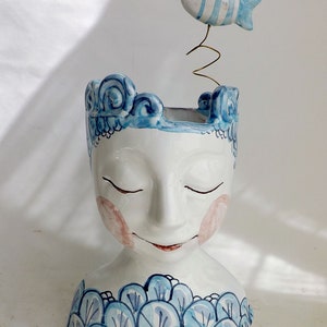 以下が含まれることがあります： 白いセラミック製の頭の形をしたプランター。目は閉じられ、頬はピンク色、口は赤く微笑んでいます。頭には青い波のような模様と、バネの上に青と白のストライプの魚が飾られています。装飾的な室内装飾。