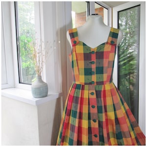 May include: A sleeveless dress featuring a vibrant plaid pattern in shades of yellow, red, green, and dark brown. The bodice has a sweetheart neckline and wide straps, with a central vertical row of dark green buttons extending down the front.