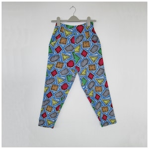 May include: Blue patterned trousers featuring a repeating design of colourful geometric shapes, including red squares, yellow triangles, and grey rectangles. The trousers are hanging on a grey hanger against a white background.