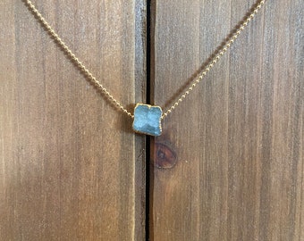 May include: A gold-colored chain necklace with a square, light blue pendant. The pendant has a rough, natural texture and is centered on the chain. The necklace is displayed against a wooden background.