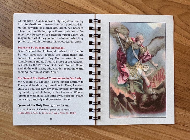 May include: Open booklet with spiral binding, featuring text and an illustration of Saint Michael the Archangel. The illustration depicts a figure with wings, holding a sword, and standing over a figure. The text includes prayers and religious information.