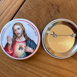 Könnte beinhalten: Ein weißer Knopf mit einem Bild von Jesus Christus mit einem roten Herzen auf seiner Brust. Der Text "Sacred Heart of Jesus" befindet sich auf der linken Seite des Knopfes und "Have Mercy on Us!" auf der rechten Seite. Die Rückseite des Knopfes ist silberfarben mit einer Stecknadel.