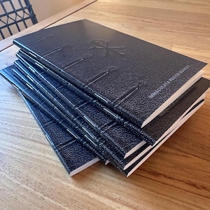 May include: A stack of five black prayer booklets with a textured cover finish. Each booklet features a debossed symbol on the front cover and the text "IMMACULATA PRAYER BOOKLET" printed in white. The booklets have visible white stitching along the spine.