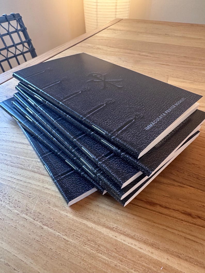 May include: A stack of five black prayer booklets with a textured cover finish. Each booklet features a debossed symbol on the front cover and the text "IMMACULATA PRAYER BOOKLET" printed in white. The booklets have visible white stitching along the spine.