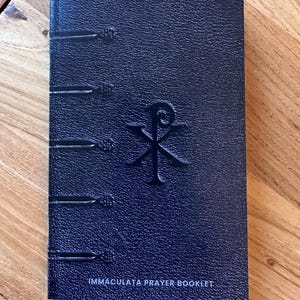 May include: A dark blue prayer booklet with a textured, leather-like cover. The front features a debossed Chi-Rho symbol in the center and five horizontal decorative bands along the left spine edge. The text IMMACULATA PRAYER BOOKLET is printed in white at the bottom.