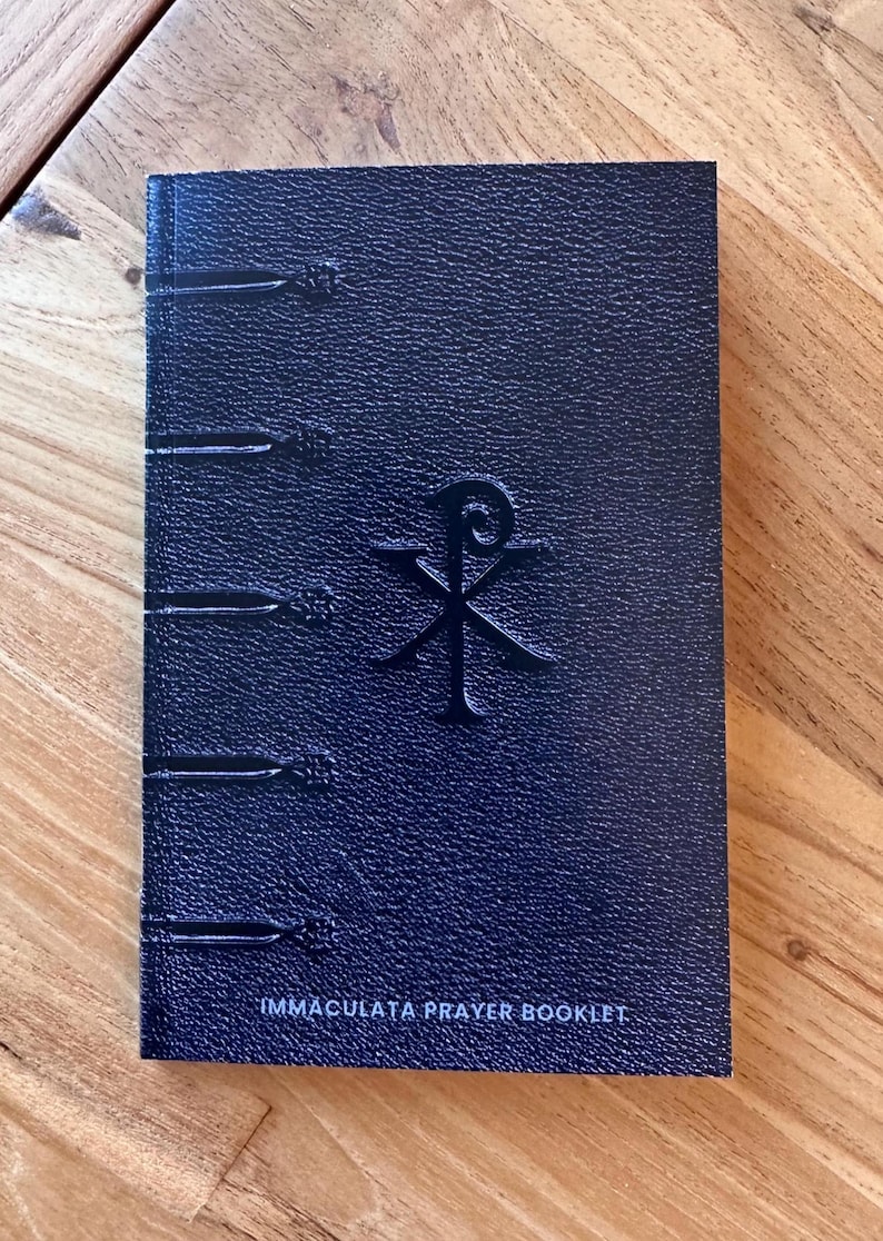 May include: A dark blue prayer booklet with a textured, leather-like cover. The front features a debossed Chi-Rho symbol in the center and five horizontal decorative bands along the left spine edge. The text IMMACULATA PRAYER BOOKLET is printed in white at the bottom.