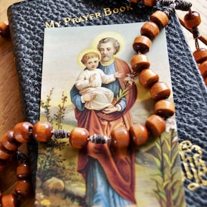 May include: A black leather prayer book with gold lettering that reads "My Prayer Book". A brown wooden rosary with a silver cord is draped over the book. A religious card with a picture of Saint Joseph holding a baby Jesus is on top of the book.