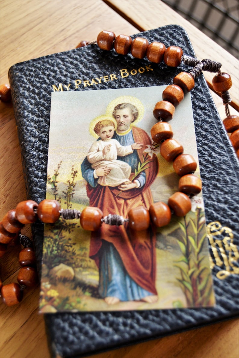 May include: A black leather prayer book with gold lettering that reads "My Prayer Book". A brown wooden rosary with a silver cord is draped over the book. A religious card with a picture of Saint Joseph holding a baby Jesus is on top of the book.