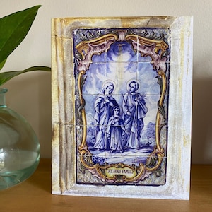 May include: A framed tile artwork depicting the Holy Family. Mary, Joseph, and baby Jesus are shown in blue robes with a gold halo. The tile is surrounded by a decorative border with gold and blue accents. The text "THE HOLY FAMILY" is written below the image.