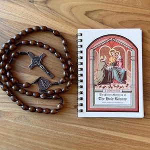 May include: A wooden rosary with brown beads and a metal crucifix and medal, next to a spiral-bound booklet. The booklet cover features an illustration of religious figures and the title "The Fifteen Mysteries of The Holy Rosary."
