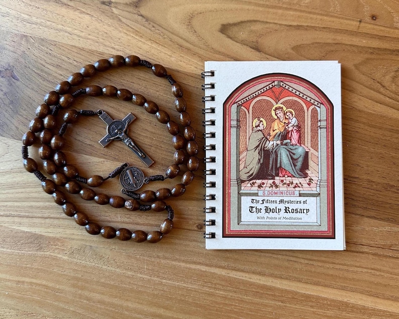 May include: A wooden rosary with brown beads and a metal crucifix and medal, next to a spiral-bound booklet. The booklet cover features an illustration of religious figures and the title "The Fifteen Mysteries of The Holy Rosary."