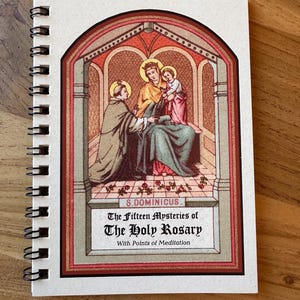 May include: A spiral-bound notebook with a religious illustration on the cover. The artwork depicts figures in robes within an arched frame, with the text "The Fifteen Mysteries of The Holy Rosary" below. The cover has a cream-colored background.