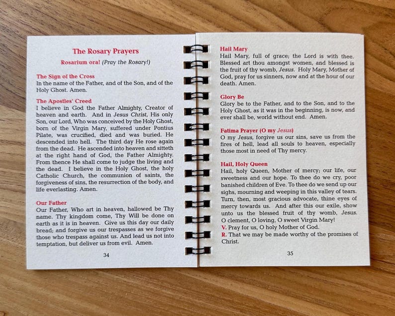 May include: Open booklet with spiral binding, featuring printed text of prayers. The left page displays "The Rosary Prayers" and "The Apostles' Creed." The right page includes "Hail Mary" and "Fatima Prayer." Pages are white with black text.