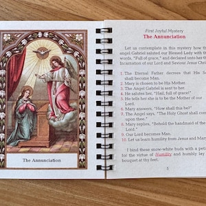 May include: A spiral-bound booklet open to a page with an illustration of The Annunciation. The artwork depicts an angel and Mary, with text describing the scene. The cover and page have a decorative border.