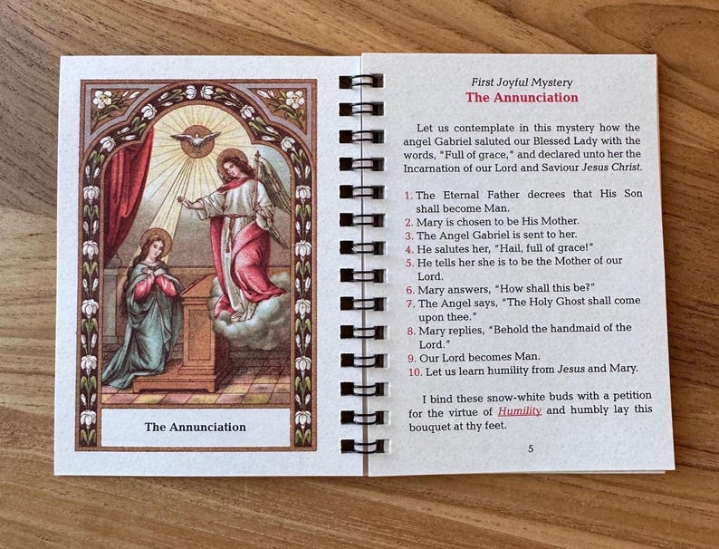 May include: A spiral-bound booklet open to a page with an illustration of The Annunciation. The artwork depicts an angel and Mary, with text describing the scene. The cover and page have a decorative border.